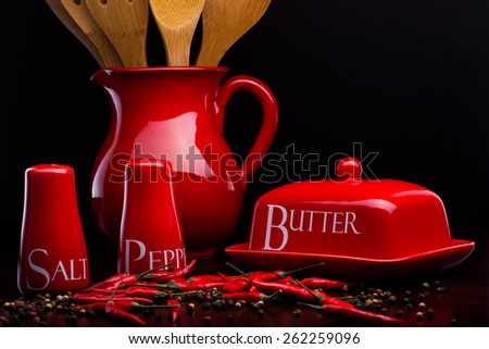 Closeup of red gloss, ceramic salt-cellar, pepper-box, butter dish and pitcher with chili and black pepper standing on dark wood table on a black background