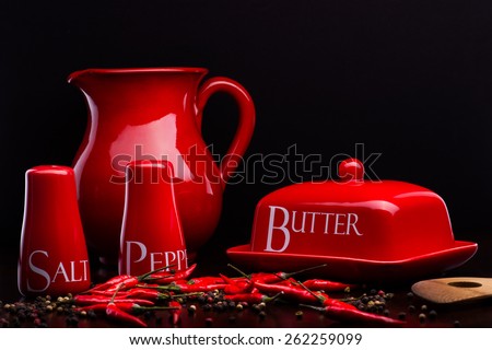 Closeup of red gloss, ceramic salt-cellar, pepper-box, butter dish and pitcher with chili and black pepper standing on dark wood table on a black background
