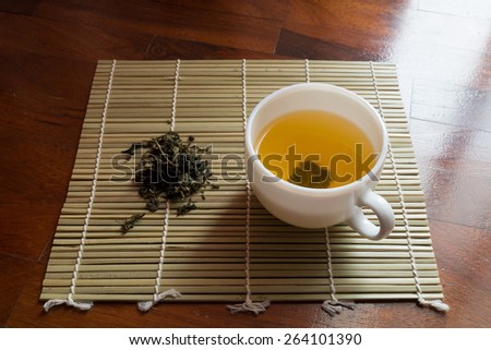 Cup of clear yellow tea and dried tea leaves on a small bamboo mat.