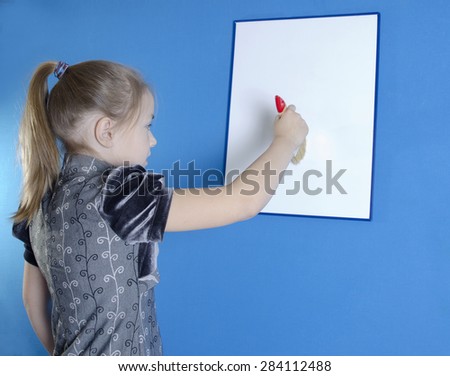 The girl of 9 years in a gray dress with black sleeves draws a big brush on a white plastic board. A photo on a blue background.