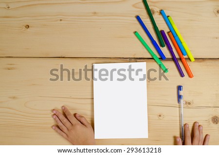 on wooden the surface lies a clean sheet of paper, nearby felt-tip pens, a pen, children\'s hands