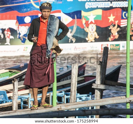 NYUANG SWHE, MYANMAR - NOVEMBER 4; Smoking Burmese man stands in bamboo path suspended over water at the docks on November 4, 2013 in Nyuang Shwe, Myanmar. Dock worker dressed in traditional longyi.