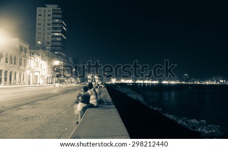 Along the Malecon, Havana, Cuba, people blurred in long exposure out at night in warm sea air sitting on sea wall across from the colonial buildings with the more modern city lights in distance.