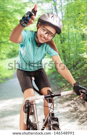 portrait of happy female sportswoman riding a race bike outside in forest, wearing professional biking gear showing thumbsup. composite image. vertical shot.