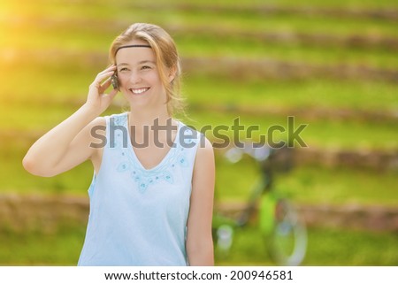 Charming Young Blond Woman Close-up Portrait in Blue Dress Speaking on Cellphone Against Green Lawn of Summer Park. Horizontal Image Composition