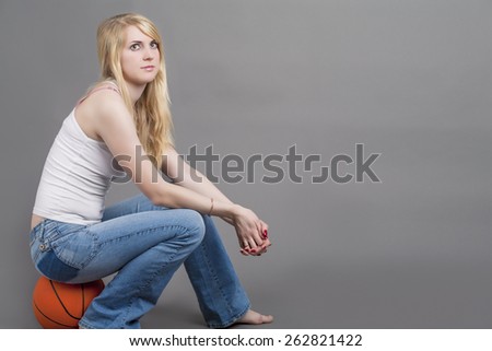 Sporty Caucasian Blond Female Sitting on Basketball Ball in Studio Environment. Horizontal Image Orientation