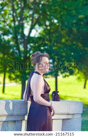 young female blonde with silk skin hands down standing turned left with head looking forward near the green trees