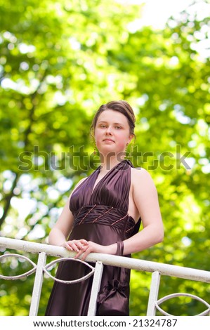 young charming lady with curly fashion hair dressing and silk skin standing on the bridge looking down body turned right smiling in sever violet dress green background