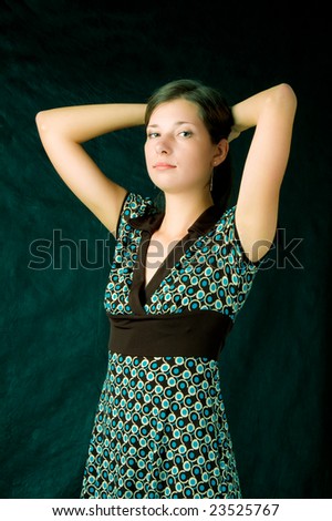 young caucasian girl with soft skin standing turned with hands up crossed behind head looking left side wearing colored dress with brown belt isolated on black