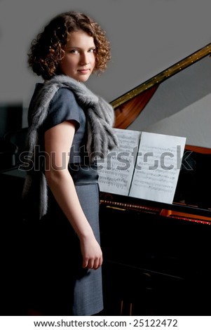 young girl with serious look in gray dress and fur scarf near black piano with music notes standing turned right looking forward on grey background