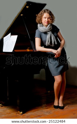 young girl with curious look in gray dress and fur scarf near black piano standing turned left supported looking forward smiling slaightly on grey background
