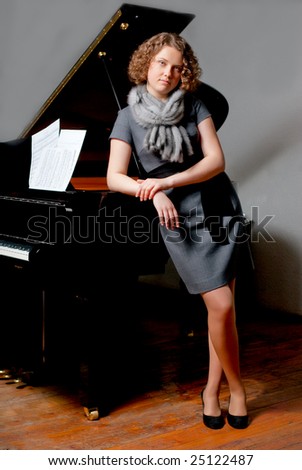 young girl with curious look in gray dress and fur scarf near black piano standing turned left supported looking forward smiling slightly on grey background