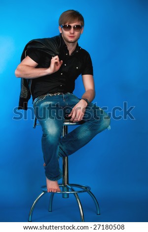 pretty young man sitting on high chair free pose looking forward in sunglasses with serious look and holding jacket ion hand behind in blue jeans,black shirt,modern watch,isolated on blue background