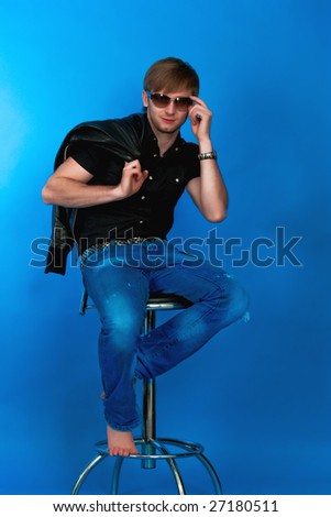 pretty young man sitting on high chair free pose looking forward touching brown sunglasses with serious look and holding jacket hand behind in blue jeans,black shirt,modern watch,isolated,background