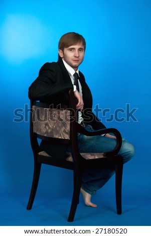 blonde man sitting on old chair turned back looking forward,wearing blue jeans,white shirt,black tie,jacket,with hands free pose appear and legs free isolated on blue