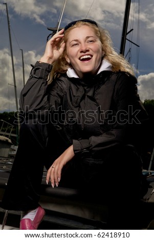 smiling caucasian blond girl on yacht sitting in waterproof jacket and holding black sunglasses. shot was made sing a flash strobe on location. high contrast.