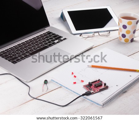 Notebook on a white table with a pen and micro controller programming kit in the middle of the background laptop, tablet, glasses and a cup
