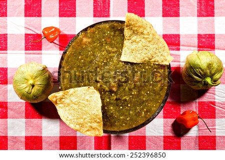 Green salsa with chips, peppers, and tomatillos on a red checkered picnic table cloth.
