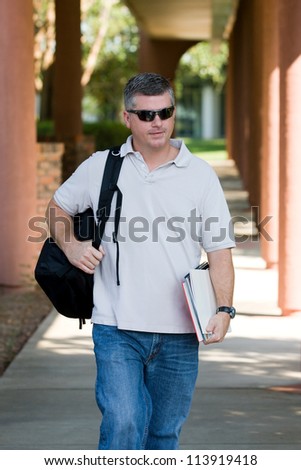 Mature adult student on campus walks to class carrying his books and backpack as he goes back to college for more education and training.