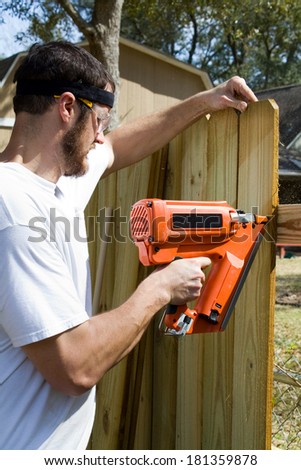 Man wearing safety glasses uses a portable nail gun to attach wood pickets to the rail as he builds a privacy fence in the backyard.