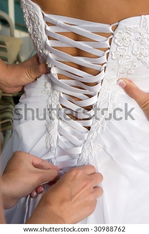 A impatient bride places hands on hips while waiting for a friend to lace up her bridal gown.