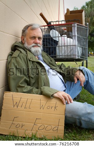 Homeless man rests leaning against a wall hold a sign with his possessions in a grocery cart.
