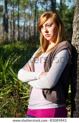 Pensive blonde female teenager with arms crossed leans her back against a tree thinking and daydreaming.