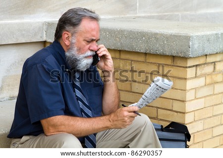Mature unemployed salesman makes a call on his cell phone to reply to a advertisement for a want ad job in the newspaper.