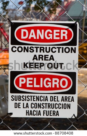 Bilingual sign in both the english and spanish language for minorities warns to keep out because of danger in a construction area is attached to fence.