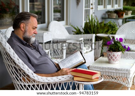 Mature man reads his bible while sitting in a white wicker chair on a porch.