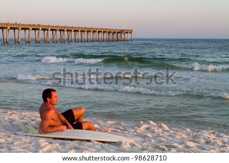 Young handsome adult surfer sits on the beach leaning on his surfboard with a pier in the background on Pensacola Beach, Florida.
