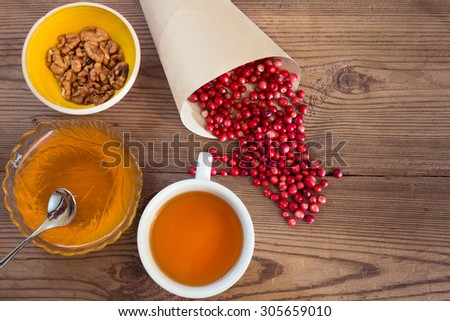 Scattered cowberries in a paper bag, honey into a bowl, walnuts in a yellow ceramic ware, white cup of tea to the left on a wooden background