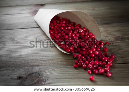 Spilling bright red cowberries in a paper bag on a wooden background.  Lots of ripe berries are poured out of a paper bag on the right background .