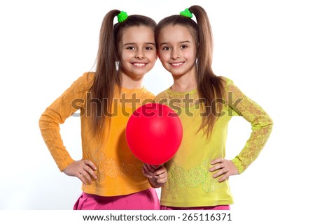 There are lovely twins with a red balloon. One girl is wearing a yellow blouse and pink trousers. Another one is wearing a green blouse and pink skirt.