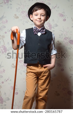 A young wizard with brown cane. A boy is wearing a light t-shirt with black waistcoat. The hat and striped bow tie complete his look