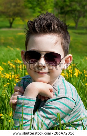 Eyewear concept. Stylish baby boy with dark hair in trendy sunglasses and in a striped polo-neck, the sitting in the park with green grass. Hipster style. Sunny weather. Close up. Outdoor shot