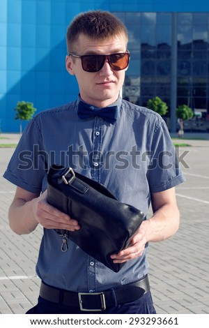 Hipster Man Concept. Portrait of attractive guy in trendy casual clothing with leather bag and sunglasses posing over shopping mall. Sunny summer weather.  Outdoor shot