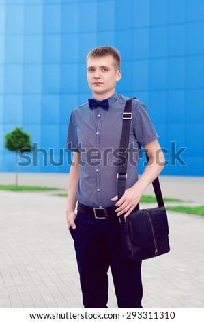 Hipster Man Concept. Portrait of attractive guy in trendy casual clothing with leather bag and  with a blue bow tie. Sunny summer weather . Outdoor shot