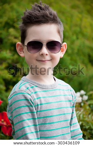 Eyewear concept. Stylish baby boy with dark hair in trendy sunglasses  and in a striped polo-neck, the  standing, in the park with green grass. Hipster style. Sunny weather. Close up. Outdoor shot