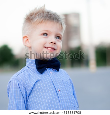 Stylish baby boy with blond hair. A boy is wearing the checked shirt and blue bow tie . He is poses on background a cities. Outdoor shot