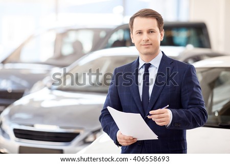 business, people, car sale and technology concept - happy smiling businessman in suit holding documents over auto show or salon background