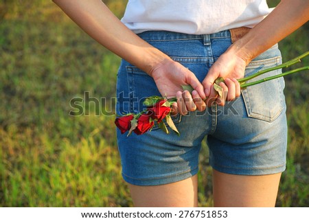 Asian young women are fall in love. She hiding the red roses behind her back and standing in the evening sunshine on the blurred meadow background.