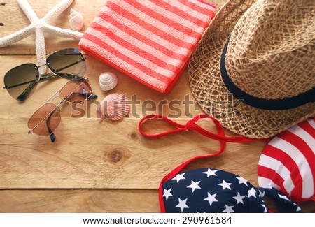 bikini is striped american flag, red striped towel, hat and sunglasses with shells and starfish are placed on a wooden background