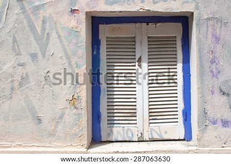 Old window with a shutter\
Wall of the old house in the part of Athens, Greece. Blue framed window with a white shutter. Under the new painting of the facade is still visible graffiti.