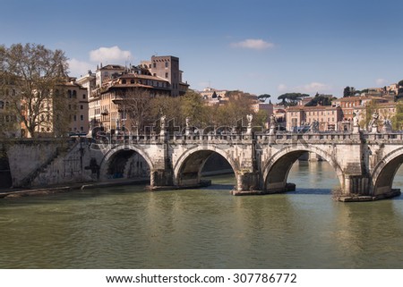 Bridge Across Tiber River, Rome, Italy\
Part of Rome close to the Castle. Old bridge over the Tiber River. Buildings of the old city in the background. Blue sky with few white clouds.