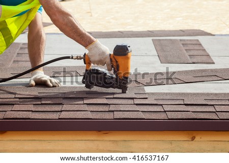 Construction worker putting the asphalt roofing (shingles) with nail gun on a new frame house