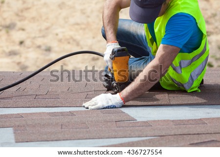 Roofer builder worker with nailgun installing Asphalt Shingles or Bitumen Tiles on a new house under construction