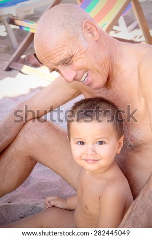 Retired grandfather and grandson in swimsuits are sitting beside the beach. The elder smiles sweetly at younger looking down and surrounding it with arms, while the child appears calm
