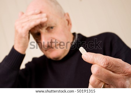 Senior older man holding a pain relief tablet or pill, while holding his head with his hand in discomfort