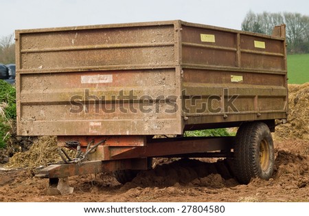 A rusty well used scruffy farm trailer parked on some manure and mud, with fields and tress in the background.